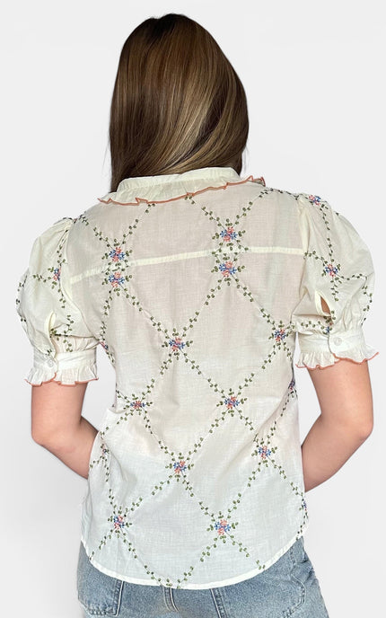 A woman viewed from the back wearing a short-sleeve cream-colored blouse with a colorful embroidered diamond pattern and light blue jeans, set against a plain light gray background.
