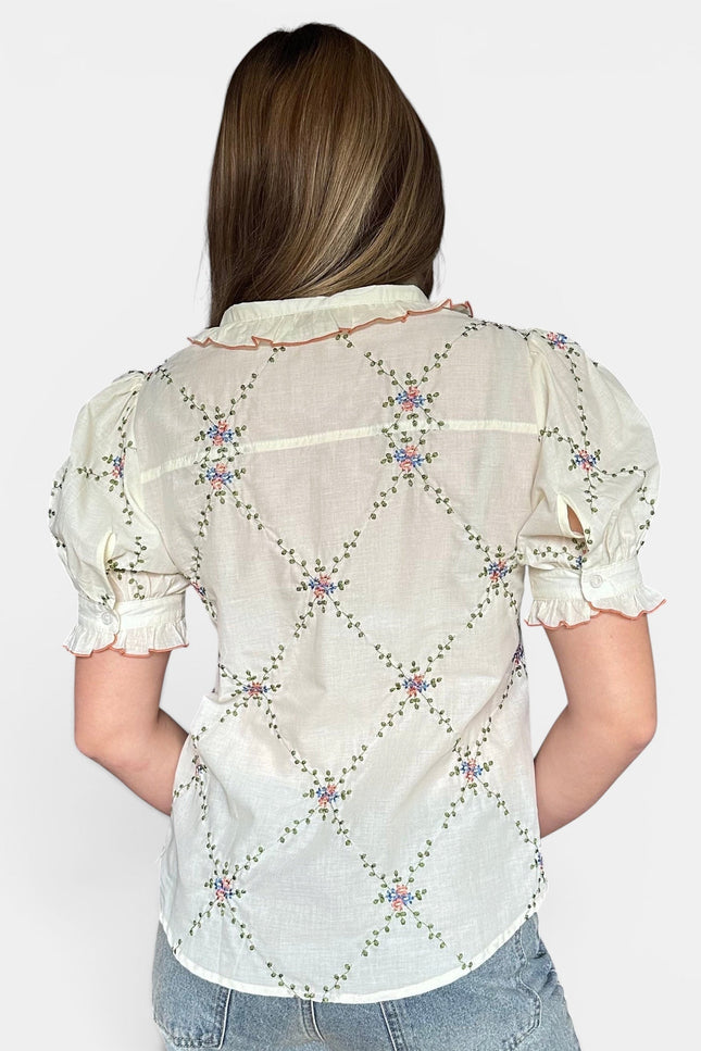 A woman viewed from the back wearing a short-sleeve cream-colored blouse with a colorful embroidered diamond pattern and light blue jeans, set against a plain light gray background.