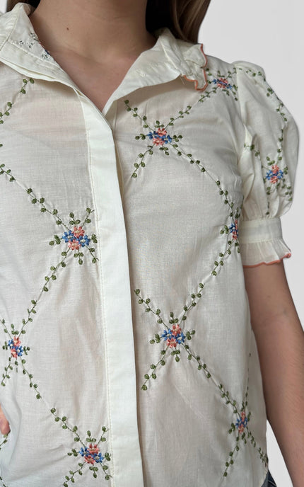 A close-up of a person wearing a cream-colored short-sleeve button-up blouse with colorful floral embroidery and green leafy vine patterns.