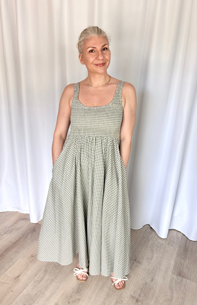 Front view of a model wearing the full-length Nora Maxi Dress in a classic gingham print, showcasing its fitted bodice, wide shoulder straps, and a gathered waist seam that flows into a long, voluminous skirt.