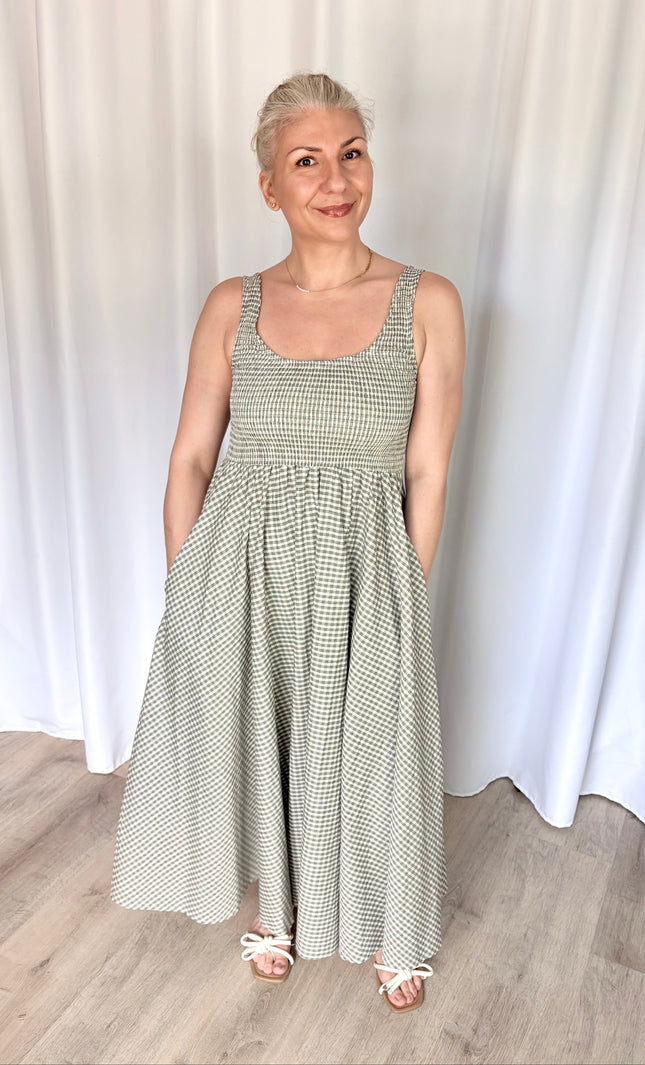 Front view of a model wearing the full-length Nora Maxi Dress in a classic gingham print, showcasing its fitted bodice, wide shoulder straps, and a gathered waist seam that flows into a long, voluminous skirt.