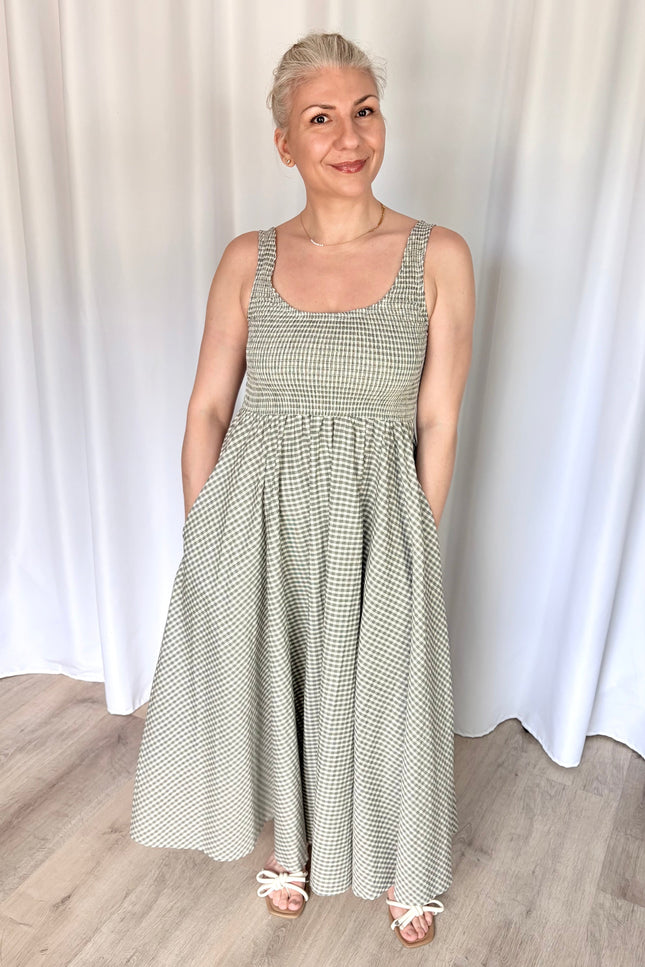 Front view of a model wearing the full-length Nora Maxi Dress in a classic gingham print, showcasing its fitted bodice, wide shoulder straps, and a gathered waist seam that flows into a long, voluminous skirt.