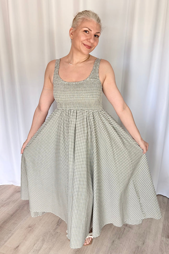 Front view of a model lightly holding the fabric of the gingham Nora Maxi Dress at each side, flaring the skirt outward to display its full, voluminous width and the graceful drape of the breathable cotton.