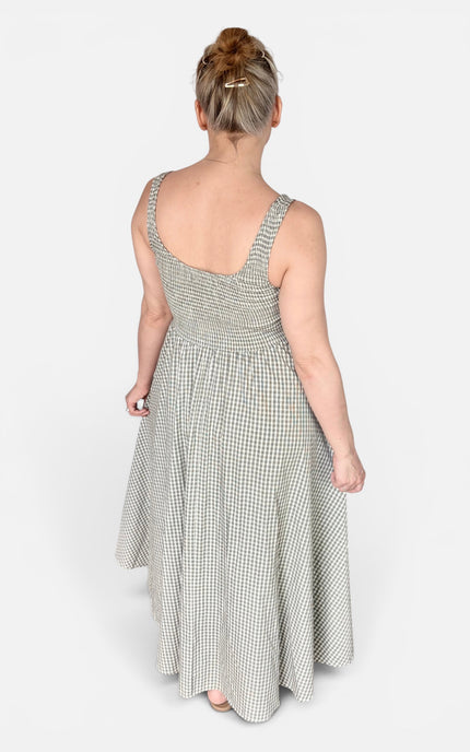 A woman viewed from behind wearing a sleeveless, smocked-waist maxi dress with a small-scale green and white gingham pattern, set against a plain light gray studio background.