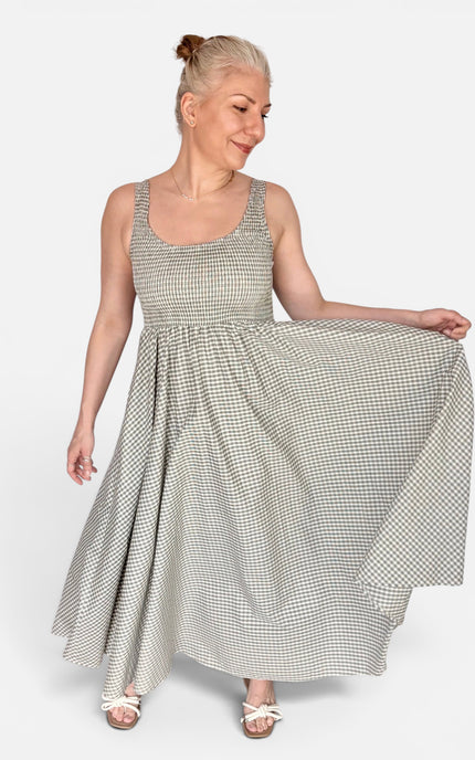 A woman modeling a sleeveless, scoop-neck maxi dress with a small-scale black and white gingham pattern, holding out the skirt to show its flow against a light gray studio background.