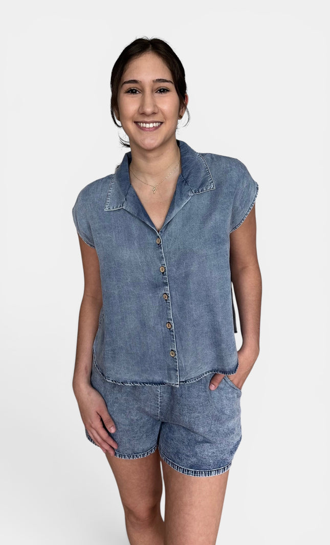 A woman modeling a light blue denim two-piece set consisting of a short-sleeve button-up shirt and matching denim shorts, shot against a plain white studio background.