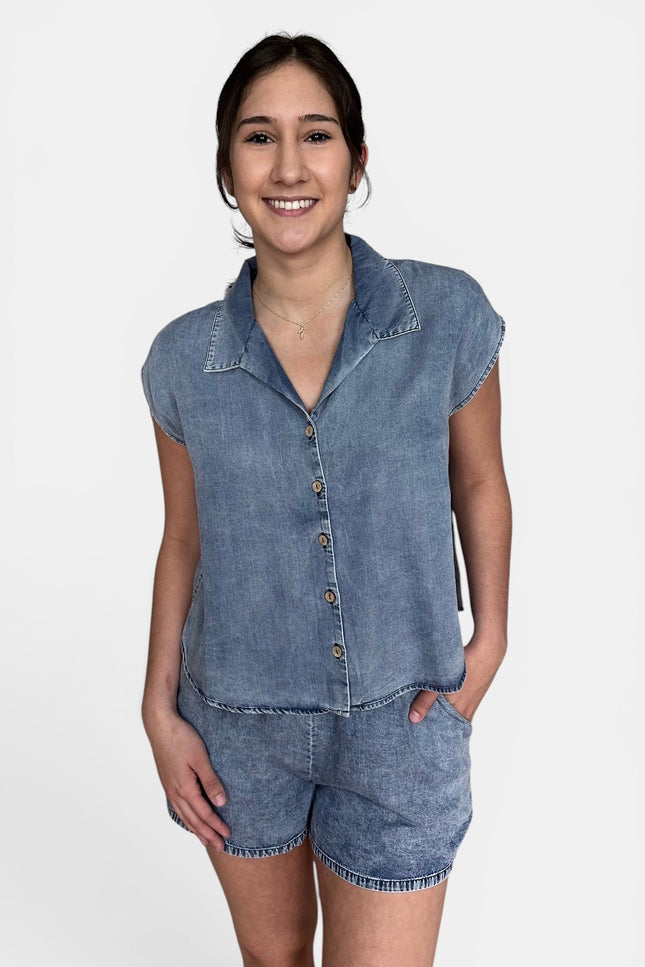 A woman modeling a light blue denim two-piece set consisting of a short-sleeve button-up shirt and matching denim shorts, shot against a plain white studio background.
