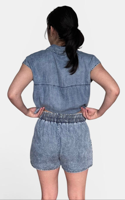 A woman viewed from behind wearing light blue tencel denim shorts paired with beige flat sandals, against a plain light gray background.
