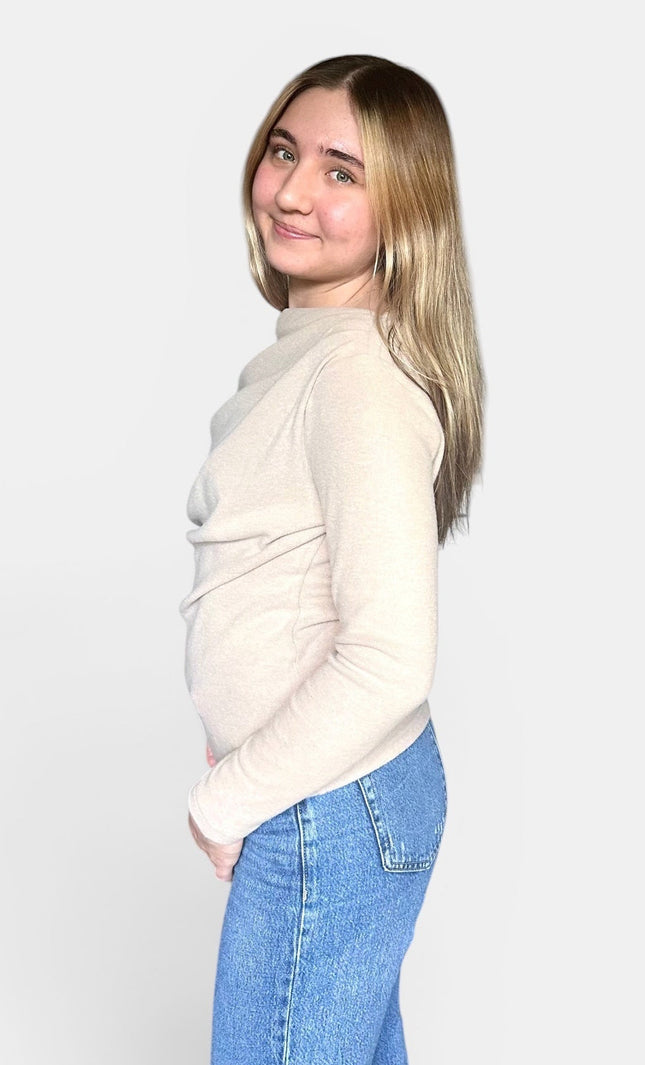A young woman with blonde hair is shown from the side profile, wearing a light beige mock-neck knit sweater and blue jeans against a plain light gray background.
