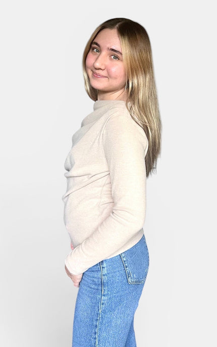 A young woman with blonde hair is shown from the side profile, wearing a light beige mock-neck knit sweater and blue jeans against a plain light gray background.