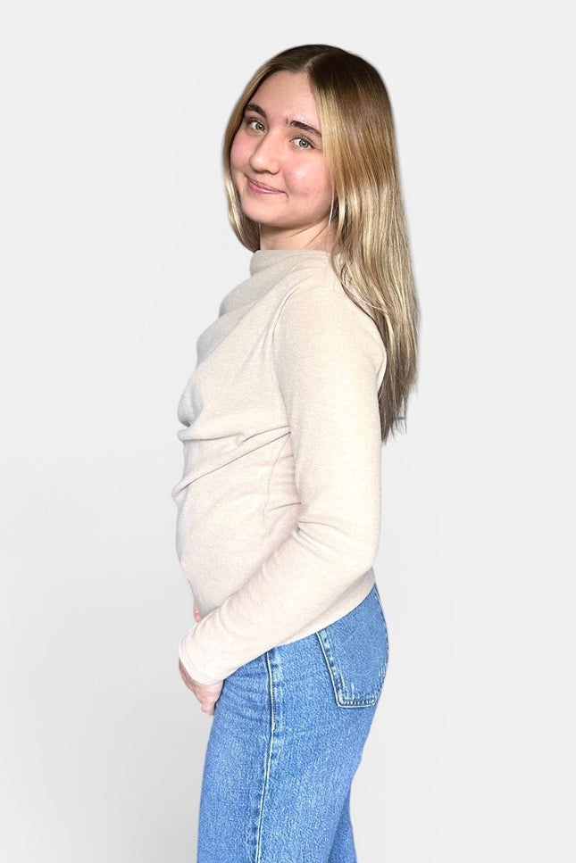A young woman with blonde hair is shown from the side profile, wearing a light beige mock-neck knit sweater and blue jeans against a plain light gray background.