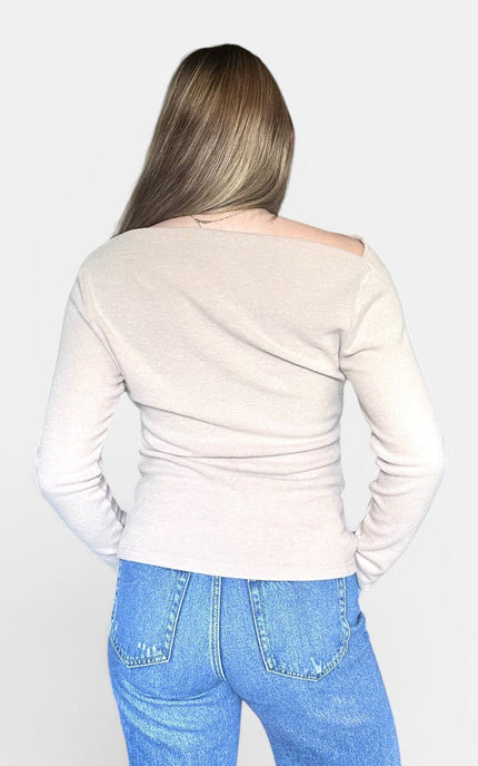 A woman viewed from behind wearing a light beige off-the-shoulder long-sleeve top and blue jeans against a plain light gray background.