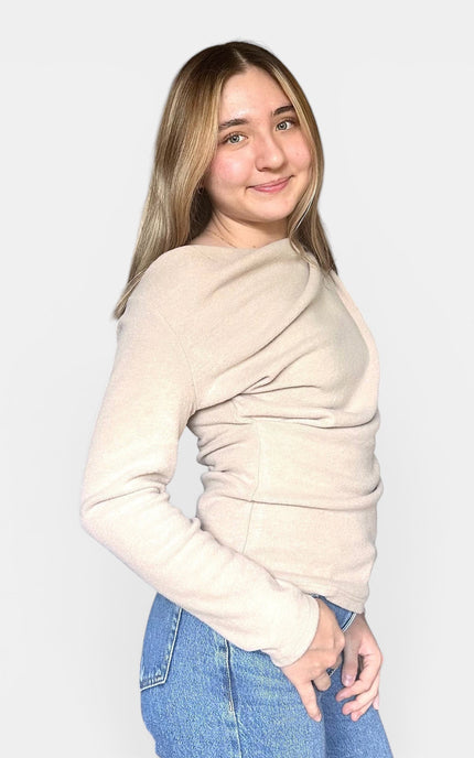 A woman with blonde hair wearing a light beige long-sleeve knit sweater and blue jeans, posing against a solid light gray background.