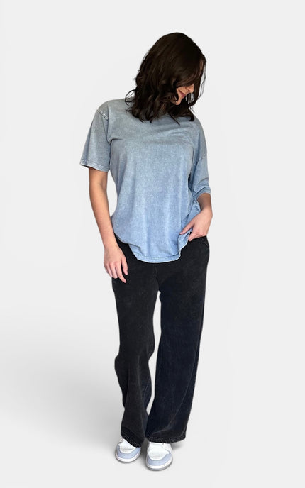 A woman wearing a light blue heathered short-sleeve t-shirt, black wide-leg pants, and white sneakers, standing against a light gray studio background.