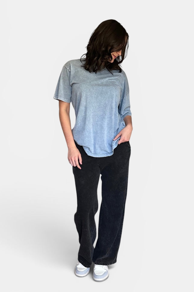 A woman wearing a light blue heathered short-sleeve t-shirt, black wide-leg pants, and white sneakers, standing against a light gray studio background.