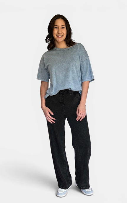 A woman modeling a light blue-gray short-sleeve t-shirt and black wide-leg jeans against a plain light gray studio background.
