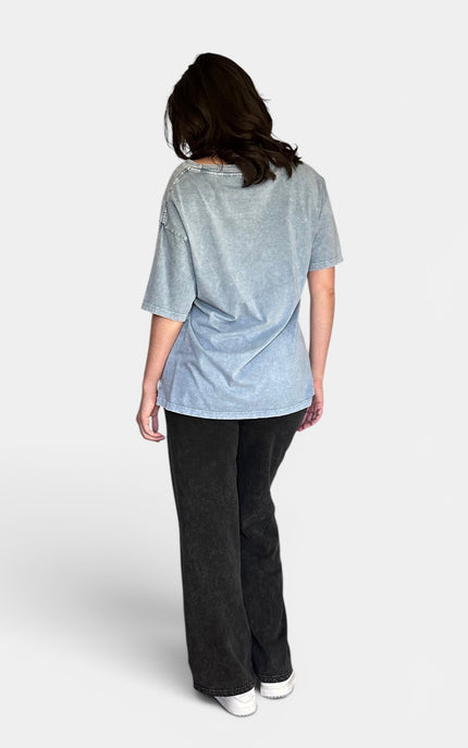 Back view of light blue-gray short-sleeve t-shirt and black wide-leg jeans against a plain light gray studio background.