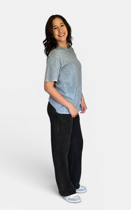 A woman modeling a light blue-gray short-sleeve t-shirt and black wide-leg pants against a plain light gray studio background.