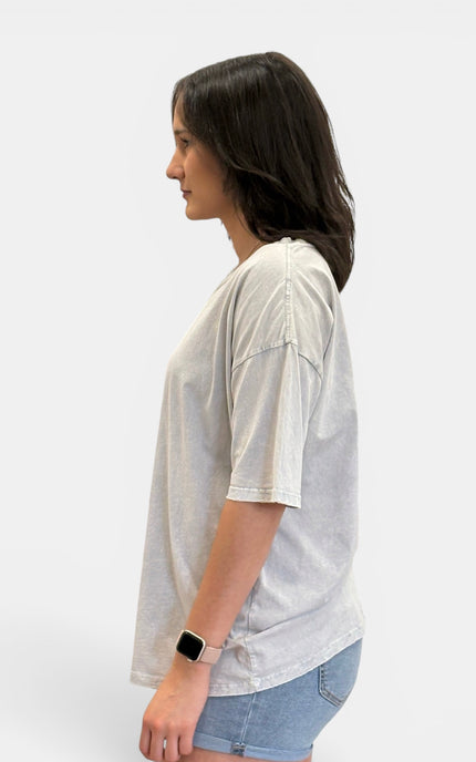 A woman shown in profile wearing a light gray oversized short-sleeve t-shirt and light blue denim shorts against a plain light gray background.