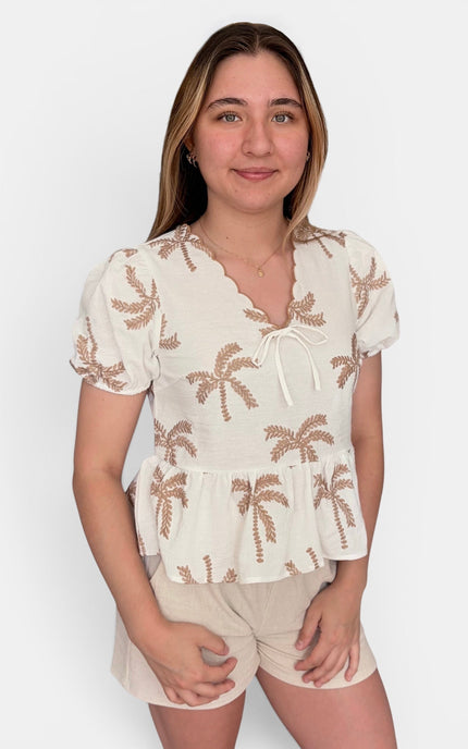 A woman modeling a short-sleeve cream top with brown palm tree embroidery and beige shorts against a plain light gray background.