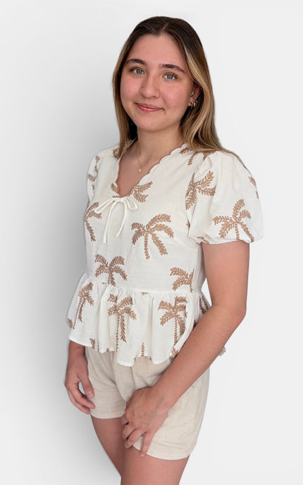 A woman modeling a short-sleeve white top with brown palm tree embroidery and matching beige shorts against a light gray studio background.