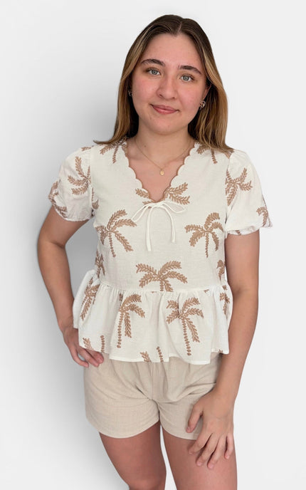 A woman modeling a short-sleeve cream blouse with brown palm tree embroidery and beige shorts against a light gray studio background.