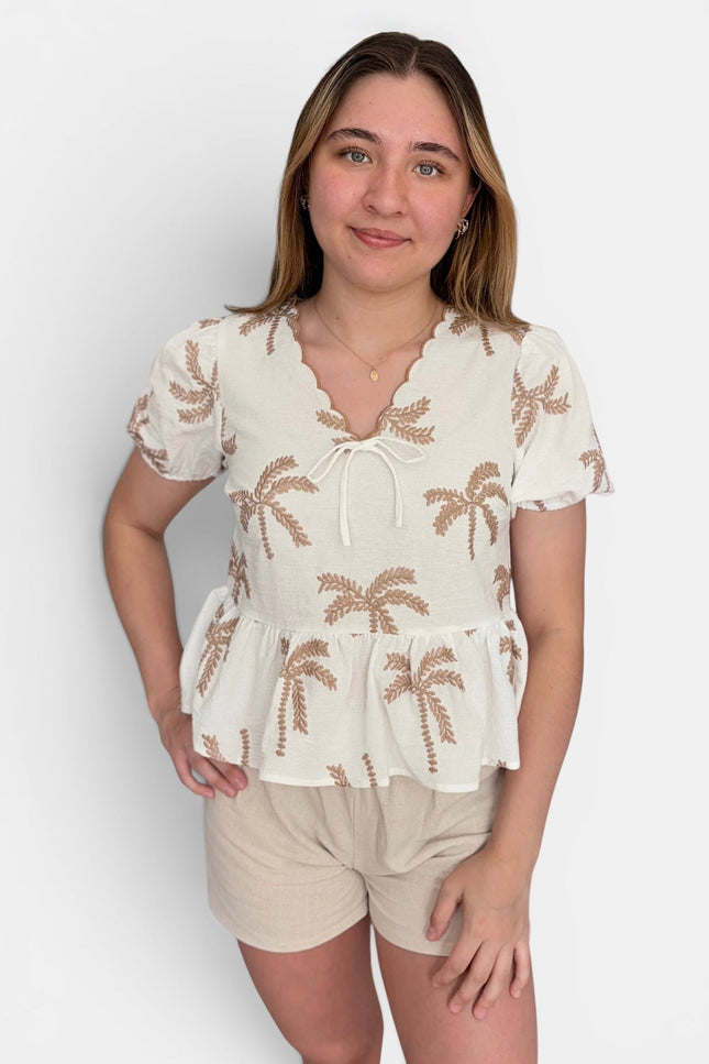 A woman modeling a short-sleeve cream blouse with brown palm tree embroidery and beige shorts against a light gray studio background.