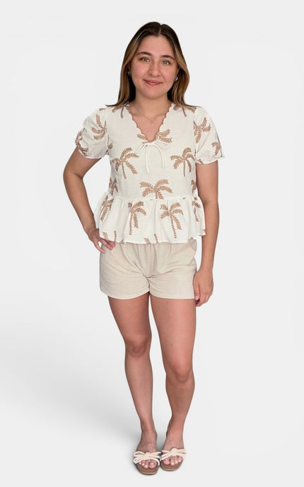 A woman modeling a two-piece casual set featuring a cream short-sleeve peplum top with a brown palm tree print and matching cream shorts, set against a plain light gray background.