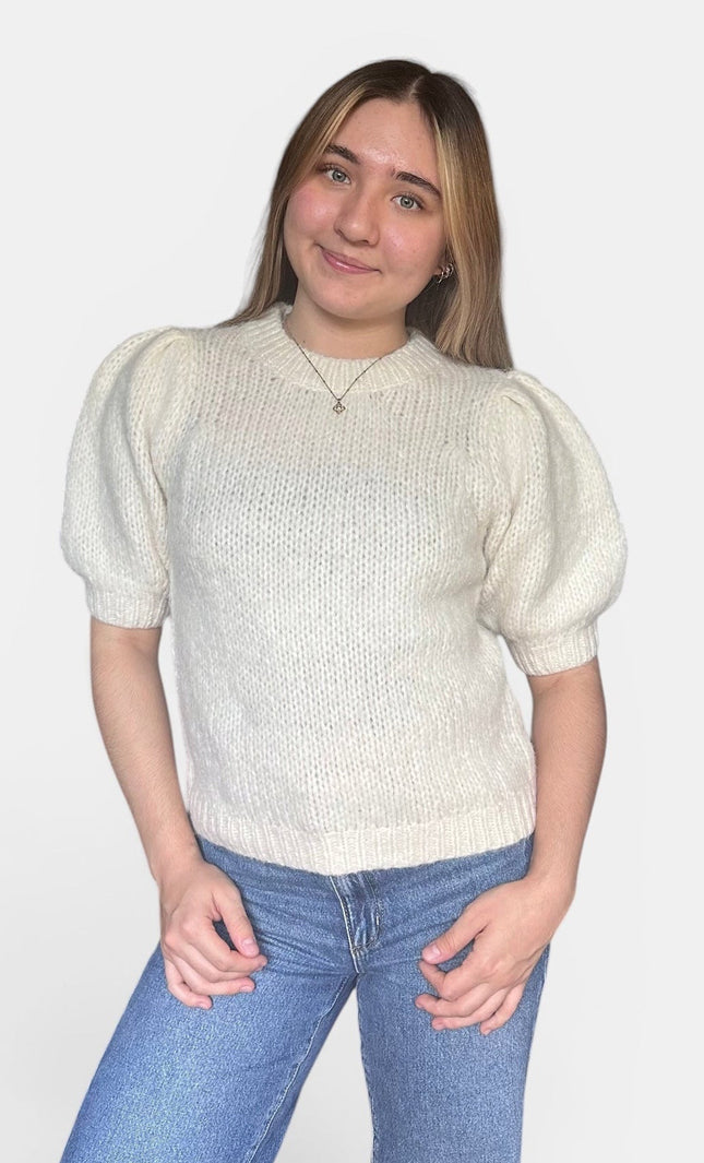 A young woman with blonde hair wearing a cream-colored short-sleeve knit sweater with puffed sleeves and blue jeans, standing against a light gray background.
