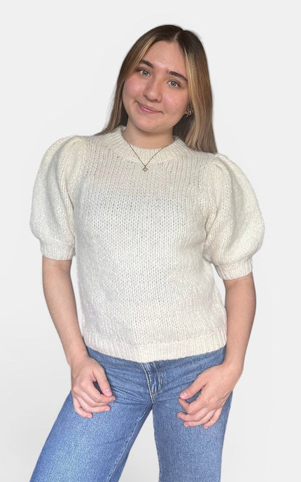 A young woman with blonde hair wearing a cream-colored short-sleeve knit sweater with puffed sleeves and blue jeans, standing against a light gray background.