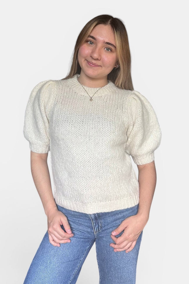 A young woman with blonde hair wearing a cream-colored short-sleeve knit sweater with puffed sleeves and blue jeans, standing against a light gray background.