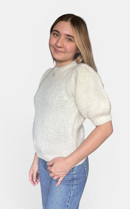 A woman modeling a cream-colored knit sweater with short puff sleeves and a crew neck, paired with blue jeans, against a plain light gray background.