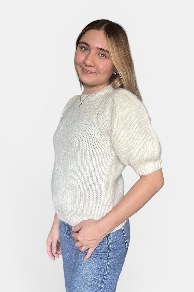 A woman modeling a cream-colored knit sweater with short puff sleeves and a crew neck, paired with blue jeans, against a plain light gray background.