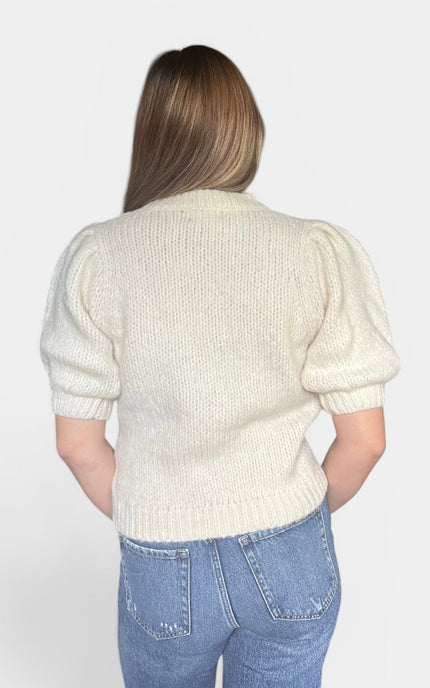 A woman viewed from the back wearing a cream-colored knit sweater with puff sleeves and blue jeans, set against a plain light gray background.