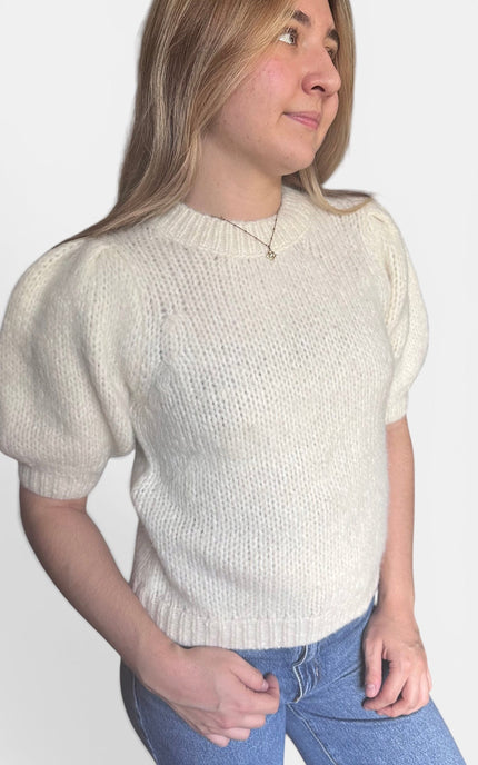 A woman wearing a cream-colored short-sleeve knit sweater with puffed shoulders and blue jeans, photographed against a plain light gray background.