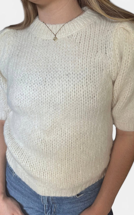 A close-up of a person wearing a cream-colored, short-sleeved knit sweater with a textured openwork pattern, paired with blue jeans.