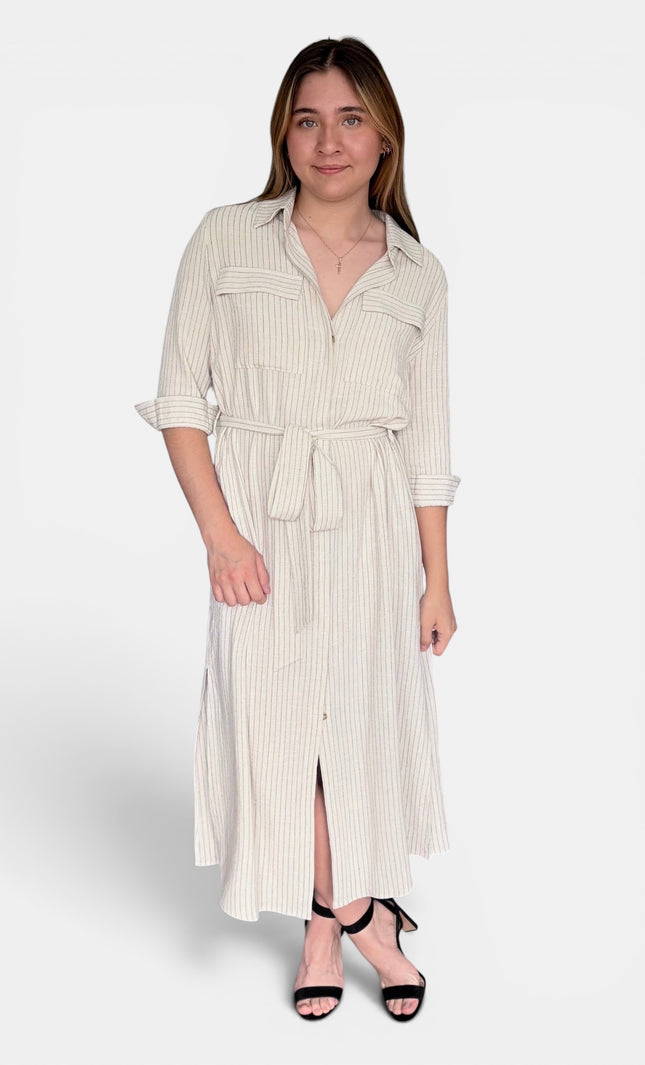 A woman modeling a cream and white vertically striped midi shirt dress with a tie waist belt, three-quarter sleeves, and a front slit, paired with black strappy heels against a light gray studio background.