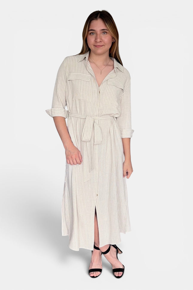 A woman modeling a cream and white vertically striped midi shirt dress with a tie waist belt, three-quarter sleeves, and a front slit, paired with black strappy heels against a light gray studio background.