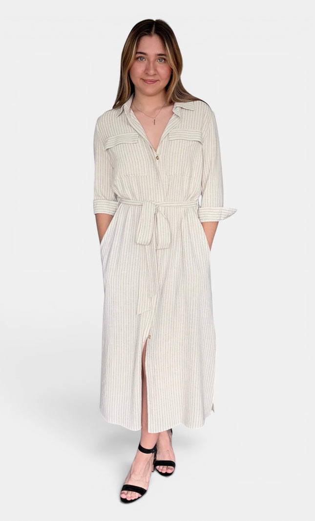 A woman modeling a cream-colored, vertically striped midi shirt dress with a tie waist and button-up front, paired with black strappy heels against a plain white studio background.