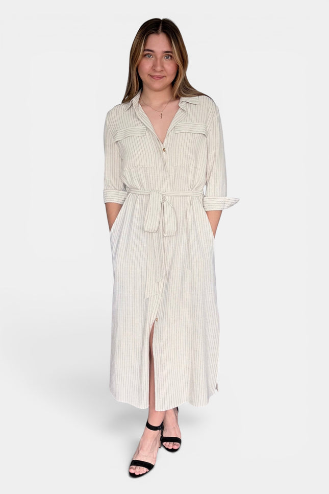 A woman modeling a cream-colored, vertically striped midi shirt dress with a tie waist and button-up front, paired with black strappy heels against a plain white studio background.