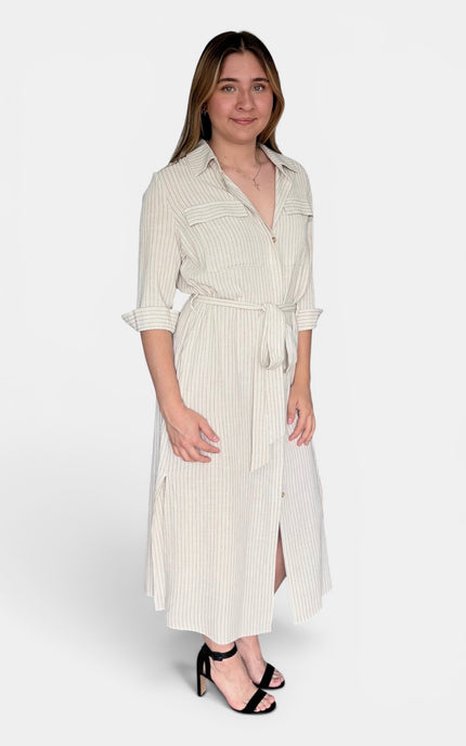 A woman modeling a cream-colored, vertically striped midi shirt dress with a tie waist belt and three-quarter sleeves, paired with black heeled sandals against a light gray studio background.