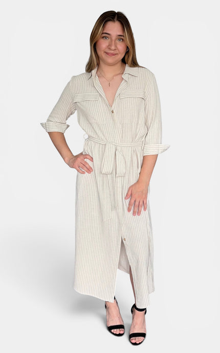 A woman modeling a long-sleeve, collared midi dress with vertical pinstripes, a self-tie waist belt, and a front slit, paired with black heeled sandals against a light gray studio background.