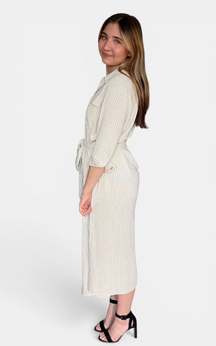 A woman modeling a cream-colored, vertically ribbed midi wrap dress with three-quarter sleeves and a tie waist, paired with black strappy heels against a light gray studio background.