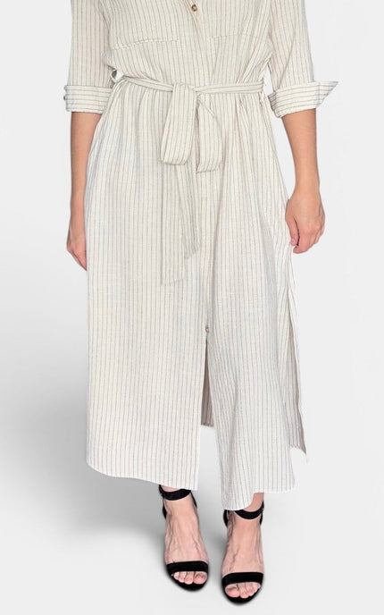 A model wearing a cream and white vertically striped wide-leg jumpsuit with a tie waist belt, paired with black strappy high-heeled sandals, against a light gray studio background.