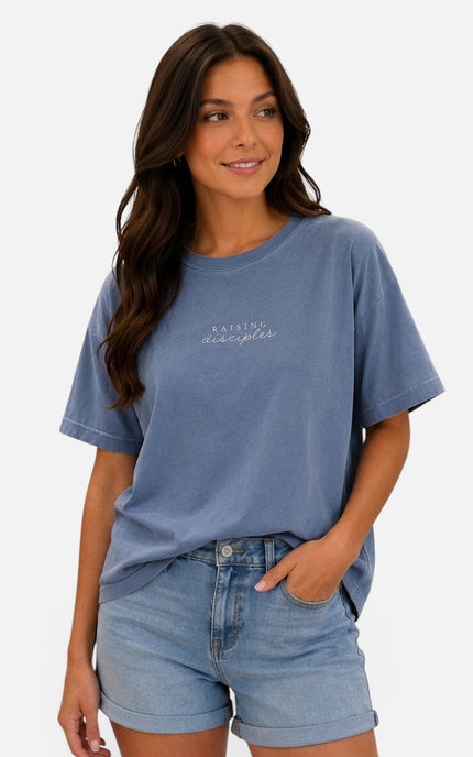 A woman wearing a faded medium blue short-sleeve t-shirt with 'Rising Despair' printed in white script on the chest, paired with light-wash denim shorts, against a plain white background.