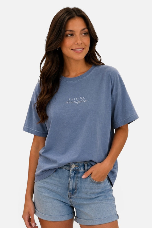 A woman wearing a faded medium blue short-sleeve t-shirt with 'Rising Despair' printed in white script on the chest, paired with light-wash denim shorts, against a plain white background.