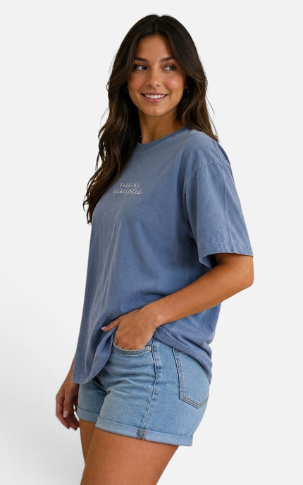 A woman wearing a faded medium blue short-sleeve t-shirt with white script text on the chest and light-wash denim shorts, posing against a solid white studio background.