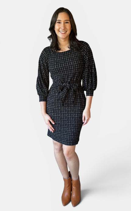 A woman modeling a black and white checkered mini dress with puff sleeves and a tie waist, paired with brown ankle boots, against a light gray studio background.