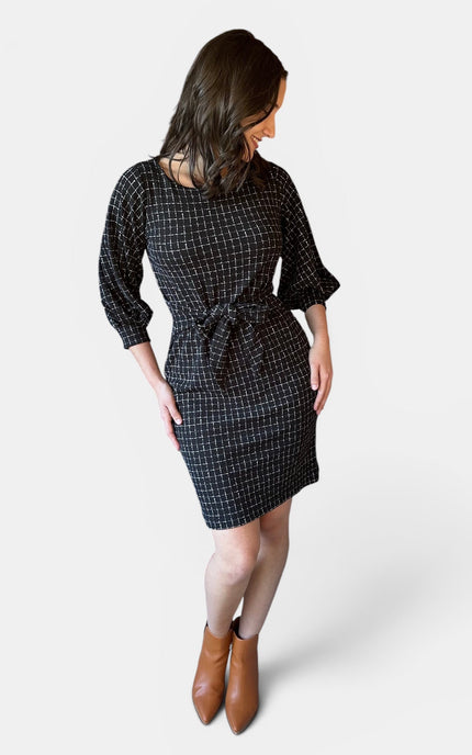 A woman modeling a black checkered mini dress with three-quarter sleeves and a tie waist, shown from a rear three-quarter angle against a light gray background.
