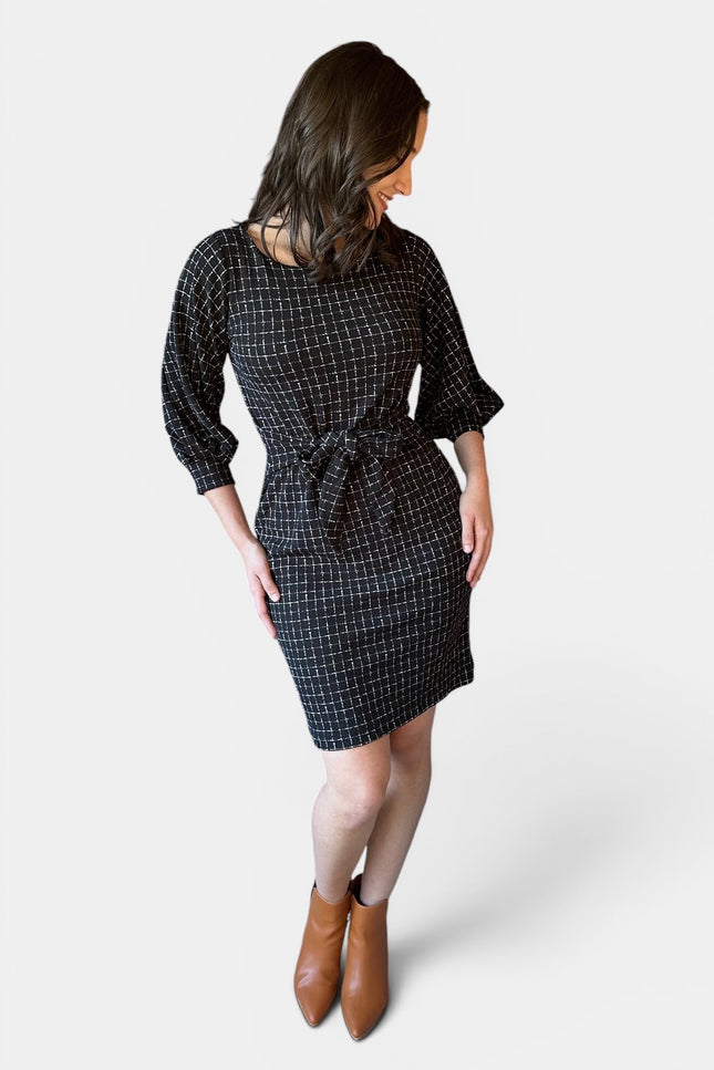 A woman modeling a black checkered mini dress with three-quarter sleeves and a tie waist, shown from a rear three-quarter angle against a light gray background.